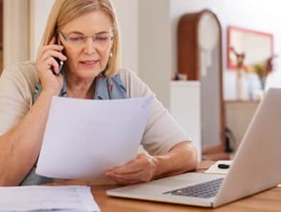An older woman on a phone talking to a laptop at home, possibly discussing massage insurance.