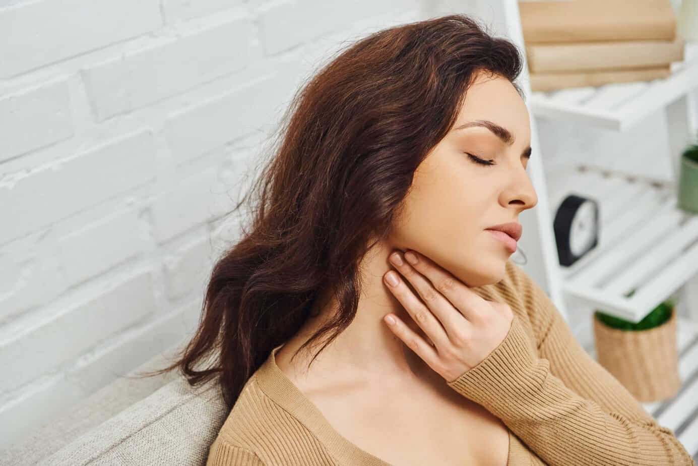 A Woman With Long Brown Hair And A Beige Sweater Is Sitting And Touching Her Neck. She Appears To Be In Discomfort And Has Her Eyes Closed. In The Background, There Is A White Brick Wall And A Shelf.