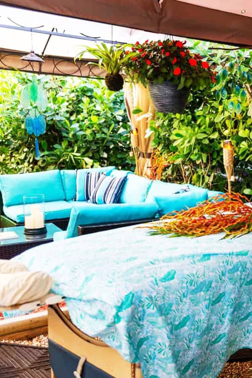 Outdoor Patio With Turquoise Furniture, Patterned Tablecloth, Hanging Plants, And Wind Chimes. Candle And Decorative Items Are Placed On Tables; Lush Green Foliage Surrounds The Patio Area.