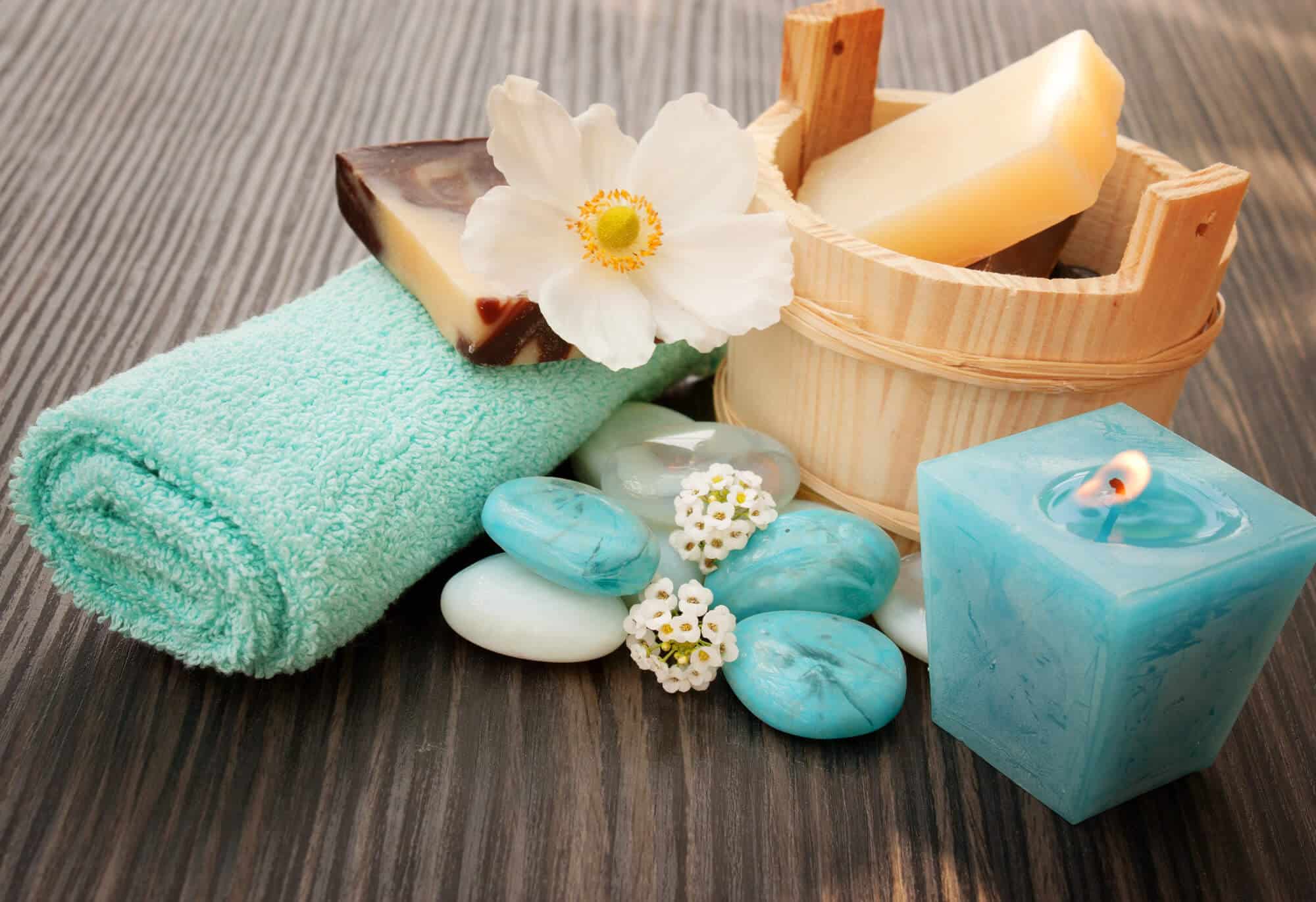 A Rolled Blue Towel, Bars Of Soap, A Wooden Container, A Lit Blue Candle, White Flowers, And Blue Stones Arranged On A Wooden Surface.