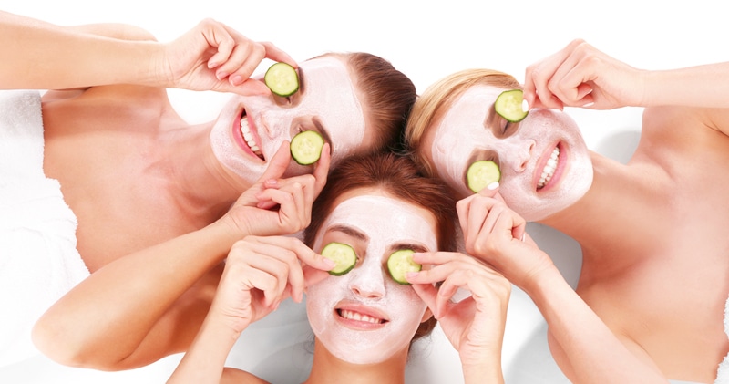 Three People With White Face Masks Lie Down, Smiling, And Hold Cucumber Slices Over Their Eyes.