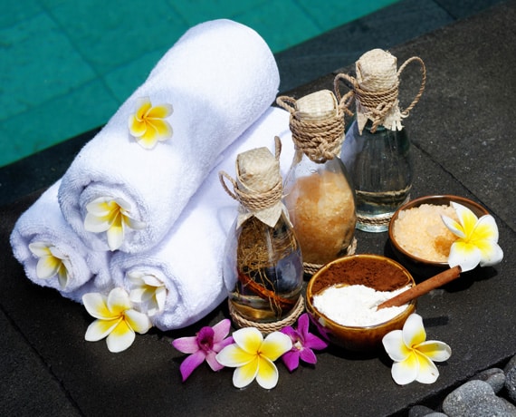 Spa Setup With Rolled White Towels, Glass Bottles With Cork Stoppers, Bowls Of Salt And Powder, Wooden Spoon, And Frangipani Flowers, Placed Near A Pool.