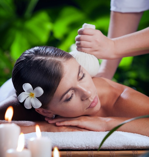 A Woman Receiving A Relaxing Massage With A Herbal Compress On Her Shoulder. Candles And Greenery In The Background.