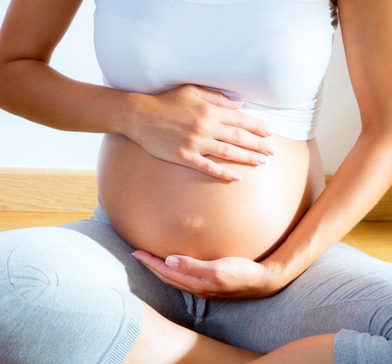 A Person Sitting On The Floor, Holding Their Pregnant Belly With Both Hands, Wearing A White Tank Top And Gray Pants.