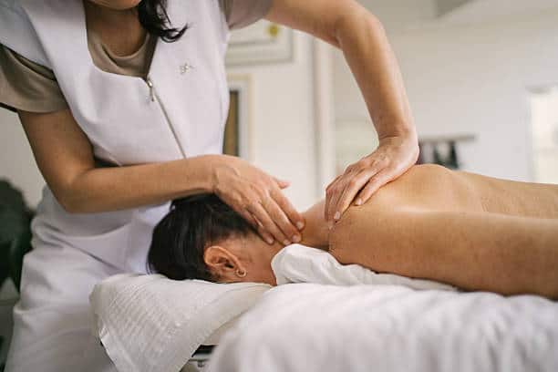 A Person Lies Face Down On A Massage Table While A Therapist Massages Their Shoulders And Neck In A Well Lit Room.