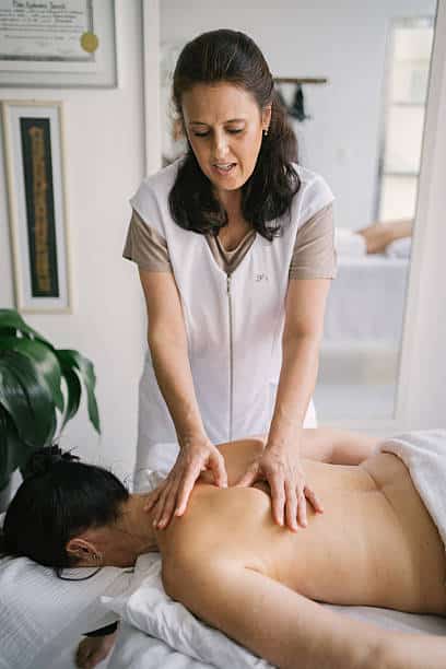 A Massage Therapist In A White Uniform Gives A Back Massage To A Person Lying Face Down On A Massage Table In A Bright Room.