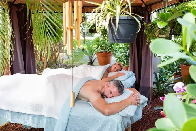Two People Lie Face Down On Massage Tables Under An Outdoor Canopy Surrounded By Lush Plants And Wind Chimes, Covered With White Towels.