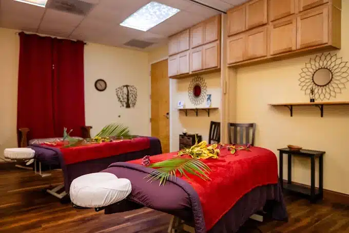 Two Massage Tables With Red Blankets And Decorative Leaves Are Set Up In A Spa Room With Wooden Cabinets, Wall Art, And Warm Lighting.