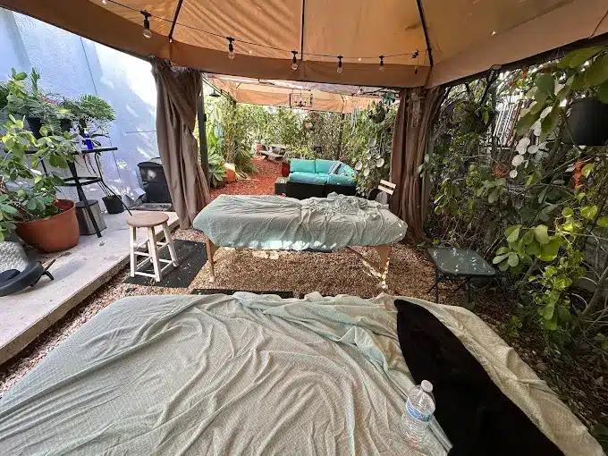Outdoor Massage Area With Shaded Canopy, Three Massage Tables Covered In Sheets, String Lights, Stools, Plants, And A Water Bottle On One Table.