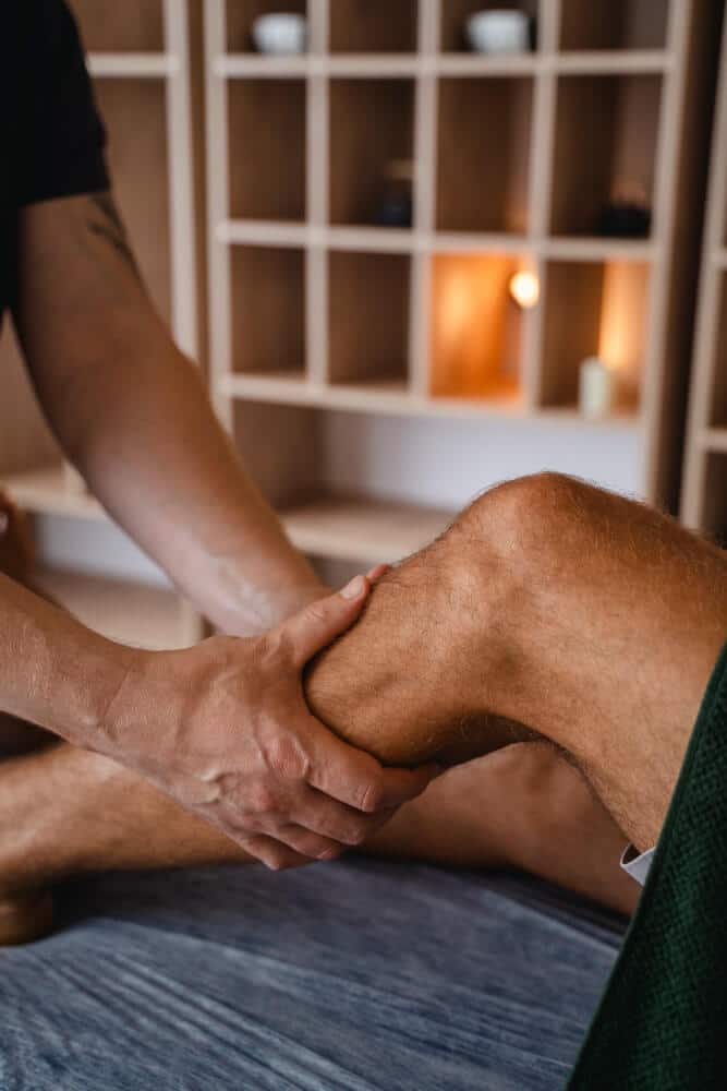A Person Receives A Knee Massage From Another Individual In A Therapy Setting With Shelves And Warm Lighting In The Background.