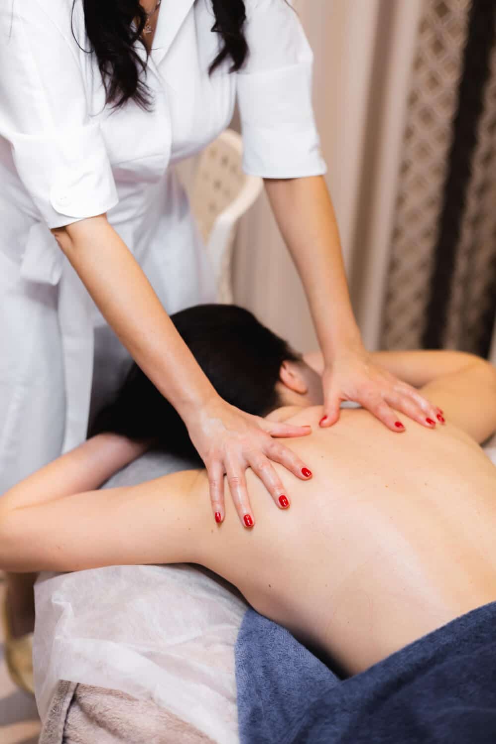 A Person Receiving A Back Massage From A Masseuse In A White Uniform; The Person Is Lying Face Down On A Massage Table Covered With A Towel.