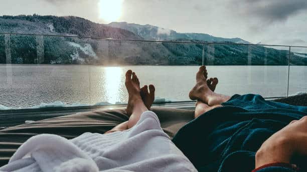 Two people in bathrobes relax on lounge chairs by a lake with snowy mountains in the background, under a cloudy sky.