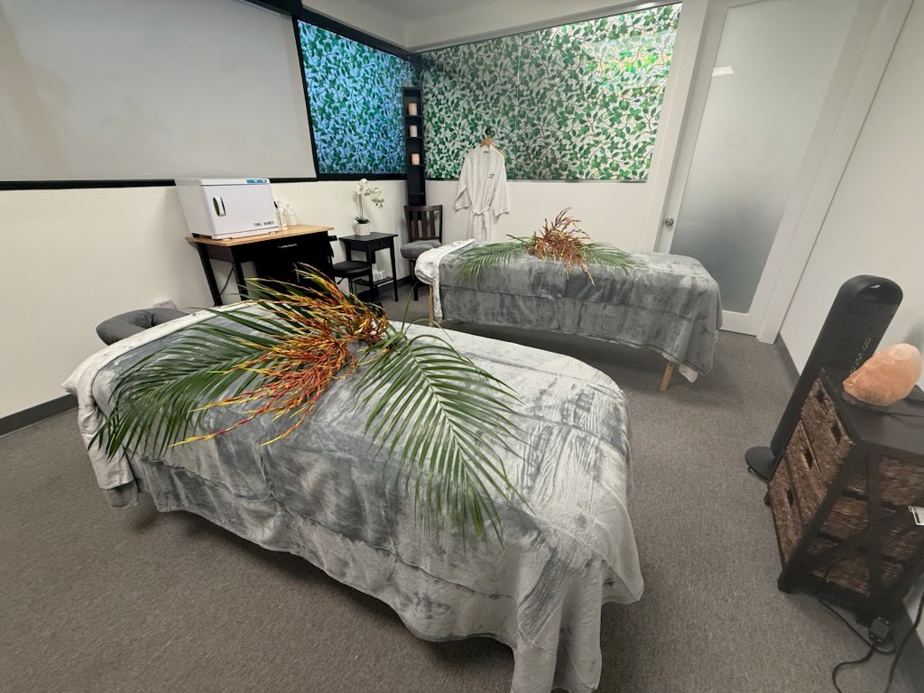 Two Massage Tables With Gray Covers, Topped With Palm Leaves And Plants, Are Set Up In A Spa Room With Green Patterned Walls And A Robe Hanging By The Door.