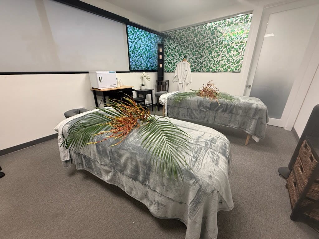 Two Massage Tables With Gray Covers And Green Leaves On Top Are Set Up In A Spa Room With White Walls, A Green Accent Wall, And A Small Desk With A Chair In The Corner.